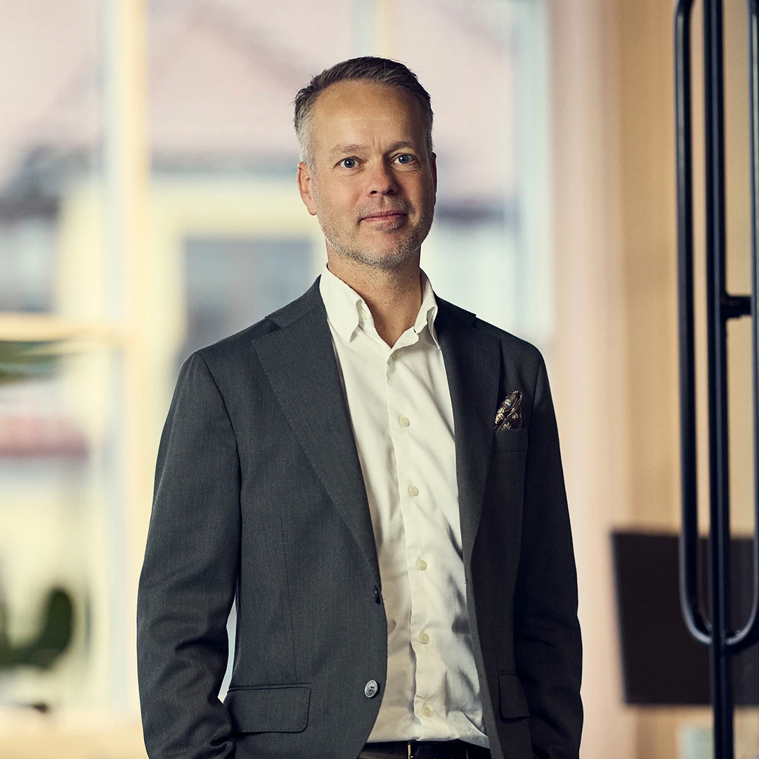 Middle-aged man in a gray suit jacket and white shirt standing indoors in a softly lit office environment.