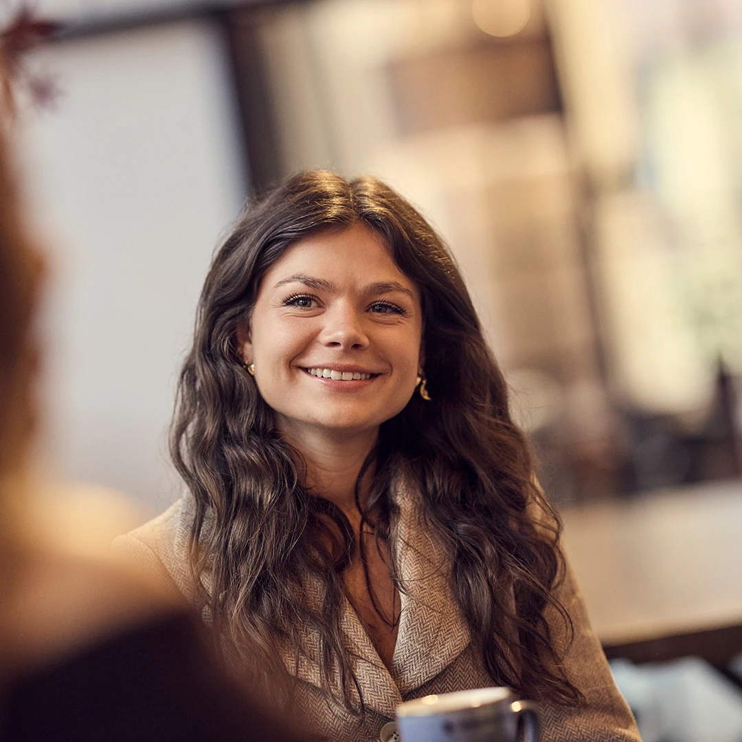 Smiling woman with long wavy hair sitting indoors holding a coffee cup.