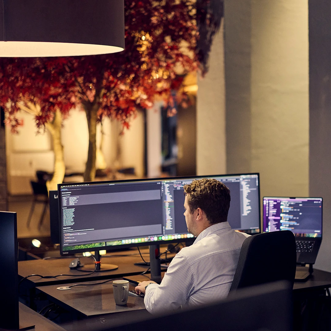 Man in a white shirt working on coding at a desk with two monitors and a mug in a dimly lit office with decorative red foliage.