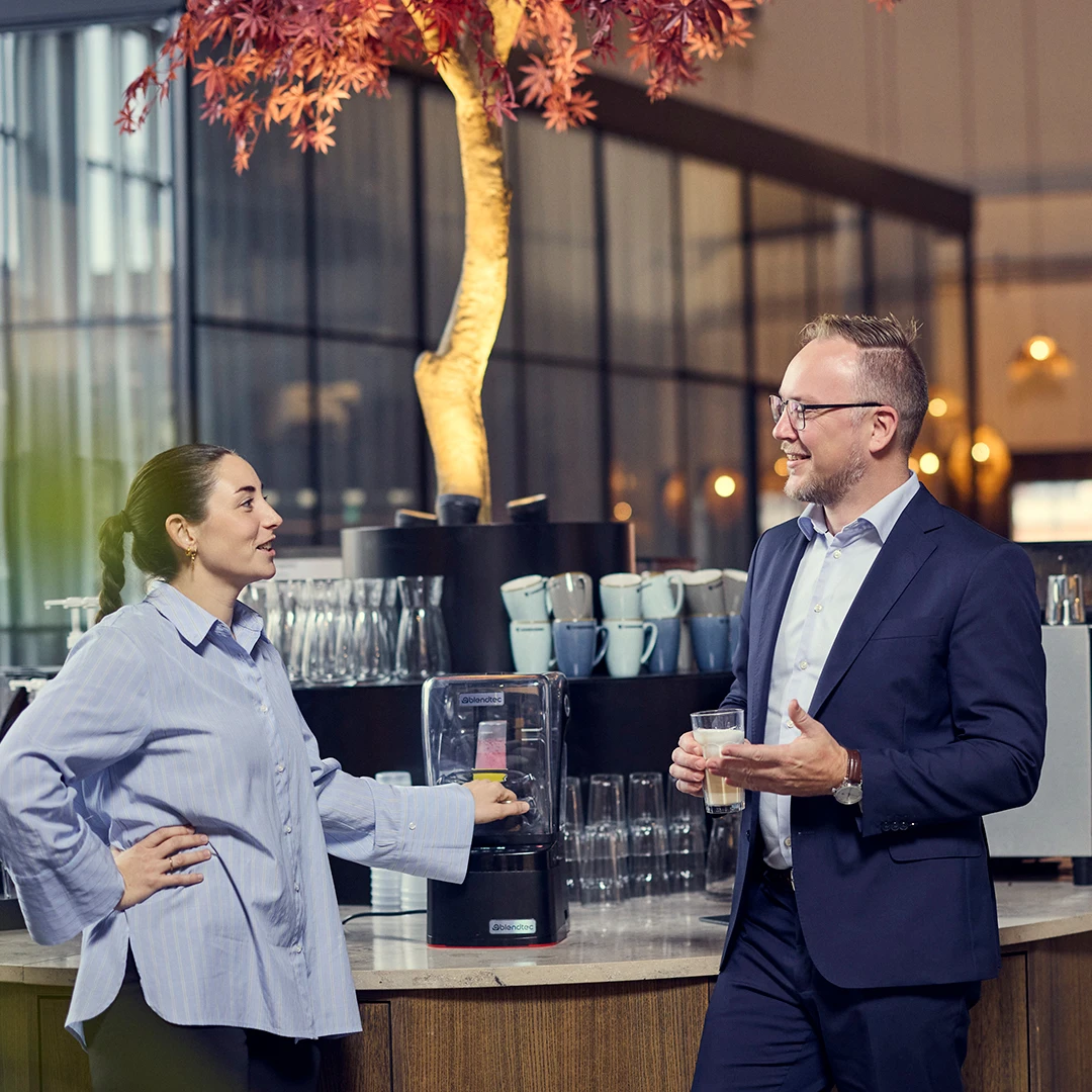 A woman and a man in business attire having a conversation in a modern office, with the woman holding a drink dispenser and the man holding a glass of coffee.