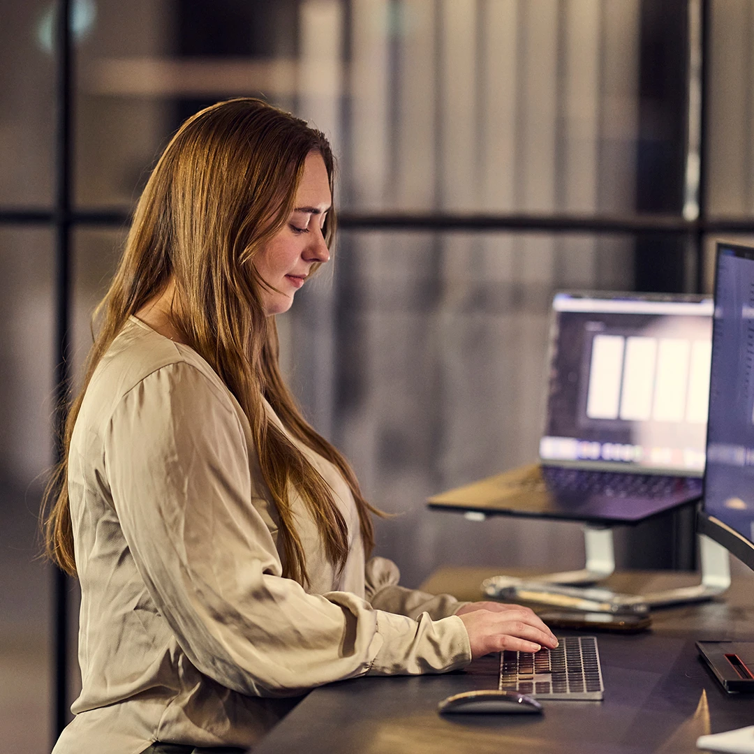 Woman with long brown hair typing on a keyboard at a desk with a laptop and monitor in the Homerunner office.
