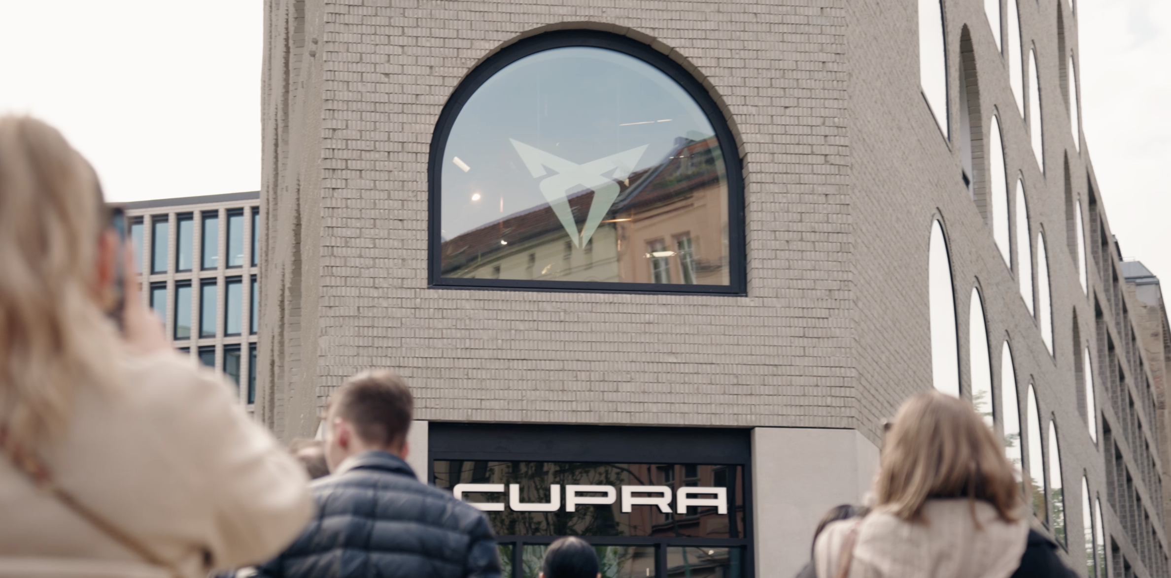 Crowd standing outside a modern brick building with large arched windows and a 'CUPRA' sign above the entrance.