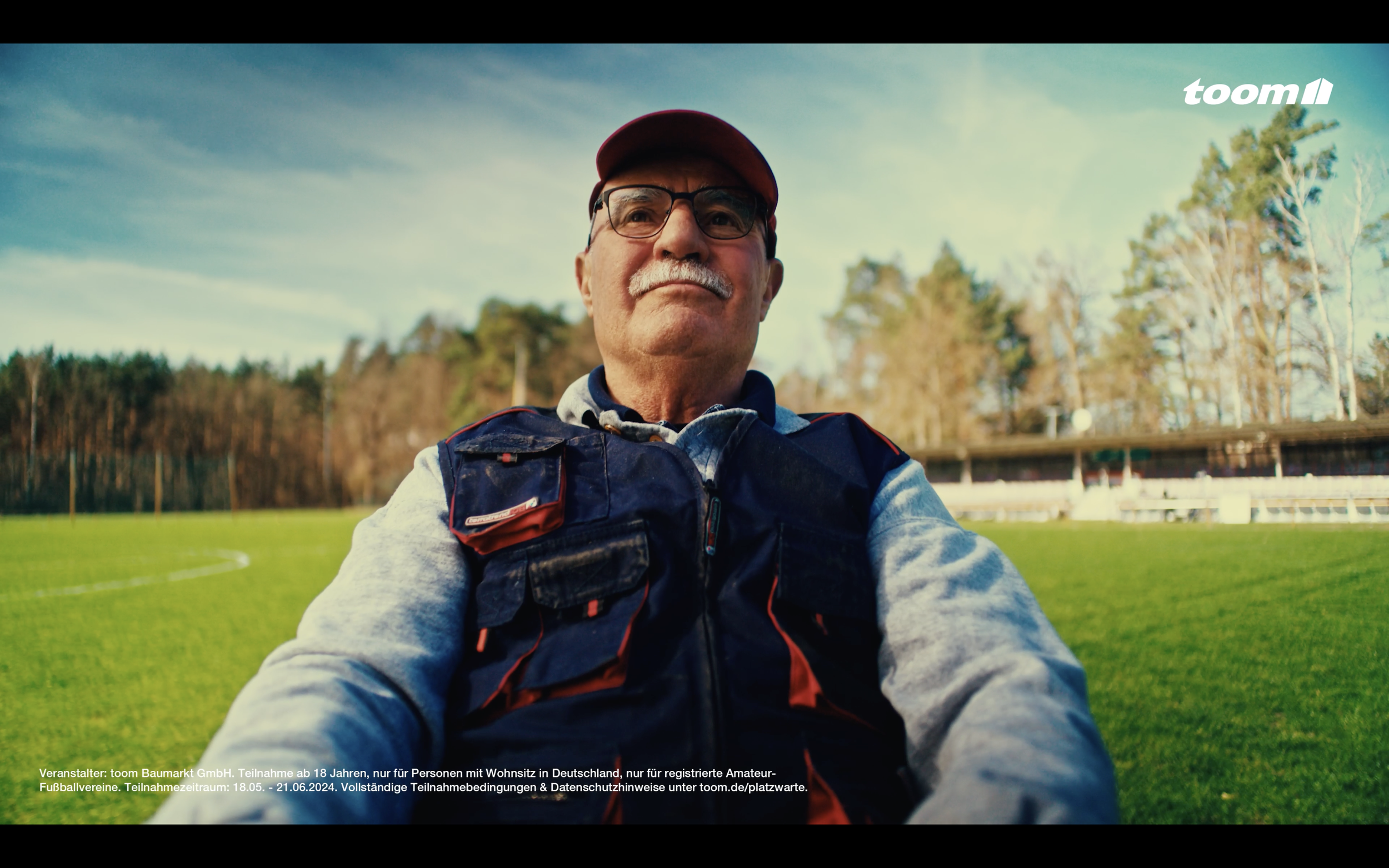 Older man wearing glasses, a red cap, and a navy-blue vest sitting outdoors on a green field with trees and a stadium in the background under a blue sky.