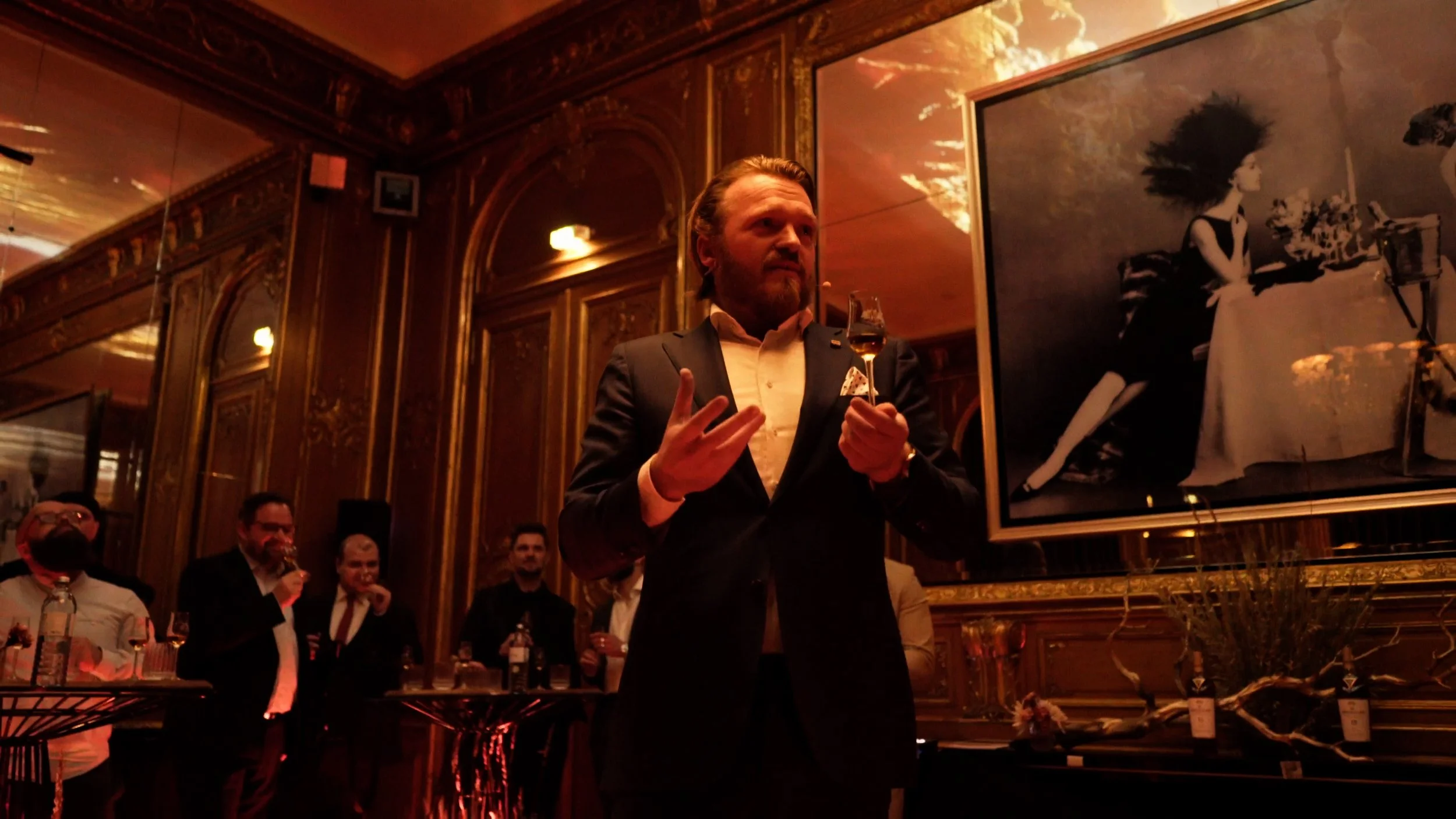Man in dark suit holding a glass of liquor speaking in a dimly lit ornate room with people and a large framed photograph in the background.