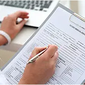 Person filling out a form on a clipboard with a pen, seated at a desk near a laptop.
