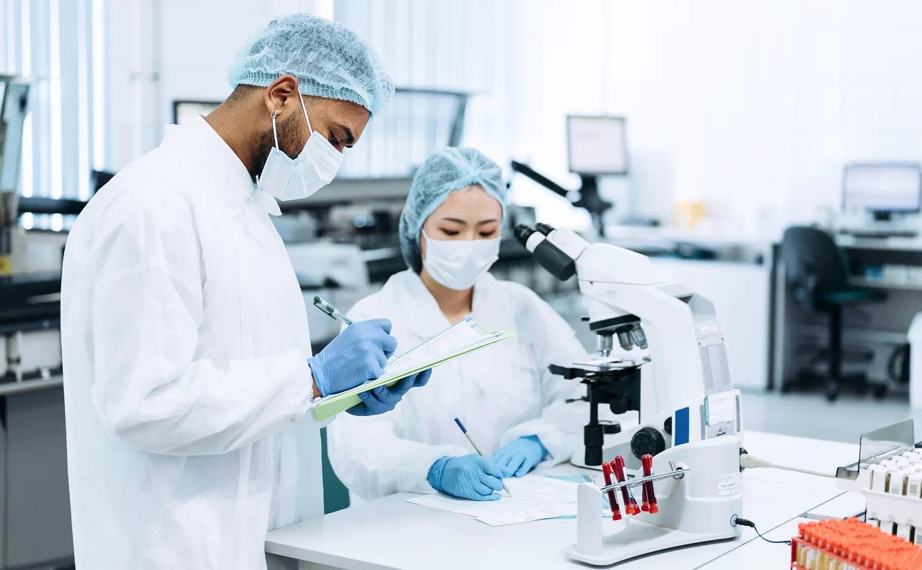 Two scientists wearing lab coats, hair covers, masks, and gloves working in a laboratory with a microscope and test tubes.