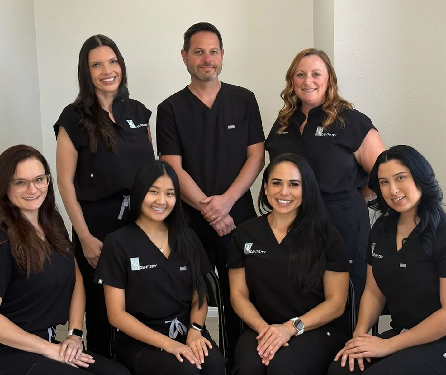 Group portrait of seven smiling dental professionals wearing black scrubs with 'Elite Dentistry' logos, standing and sitting against a plain wall.