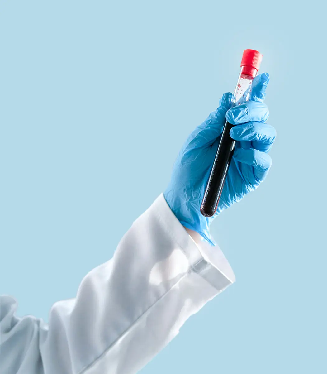 Hand in blue glove holding a test tube filled with dark red liquid against a light blue background.