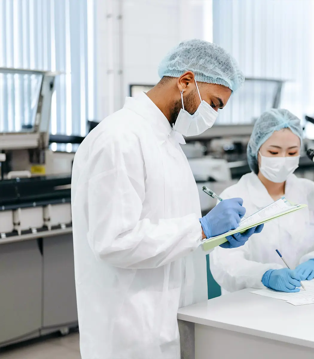Two scientists in lab coats, hairnets, and masks working in a laboratory, one writing on a clipboard and the other taking notes.