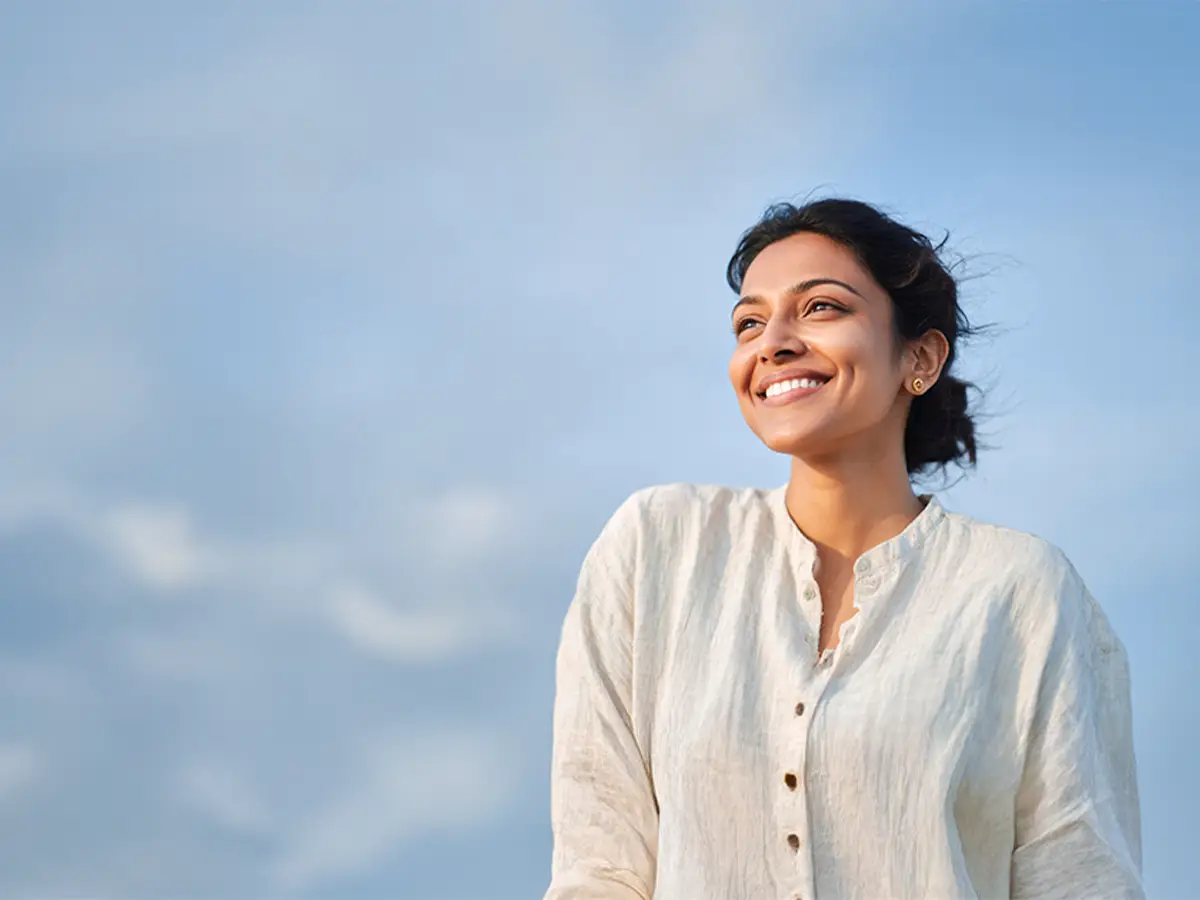 Smiling woman with dark hair in a light beige buttoned shirt looking to the right against a blue sky backdrop.