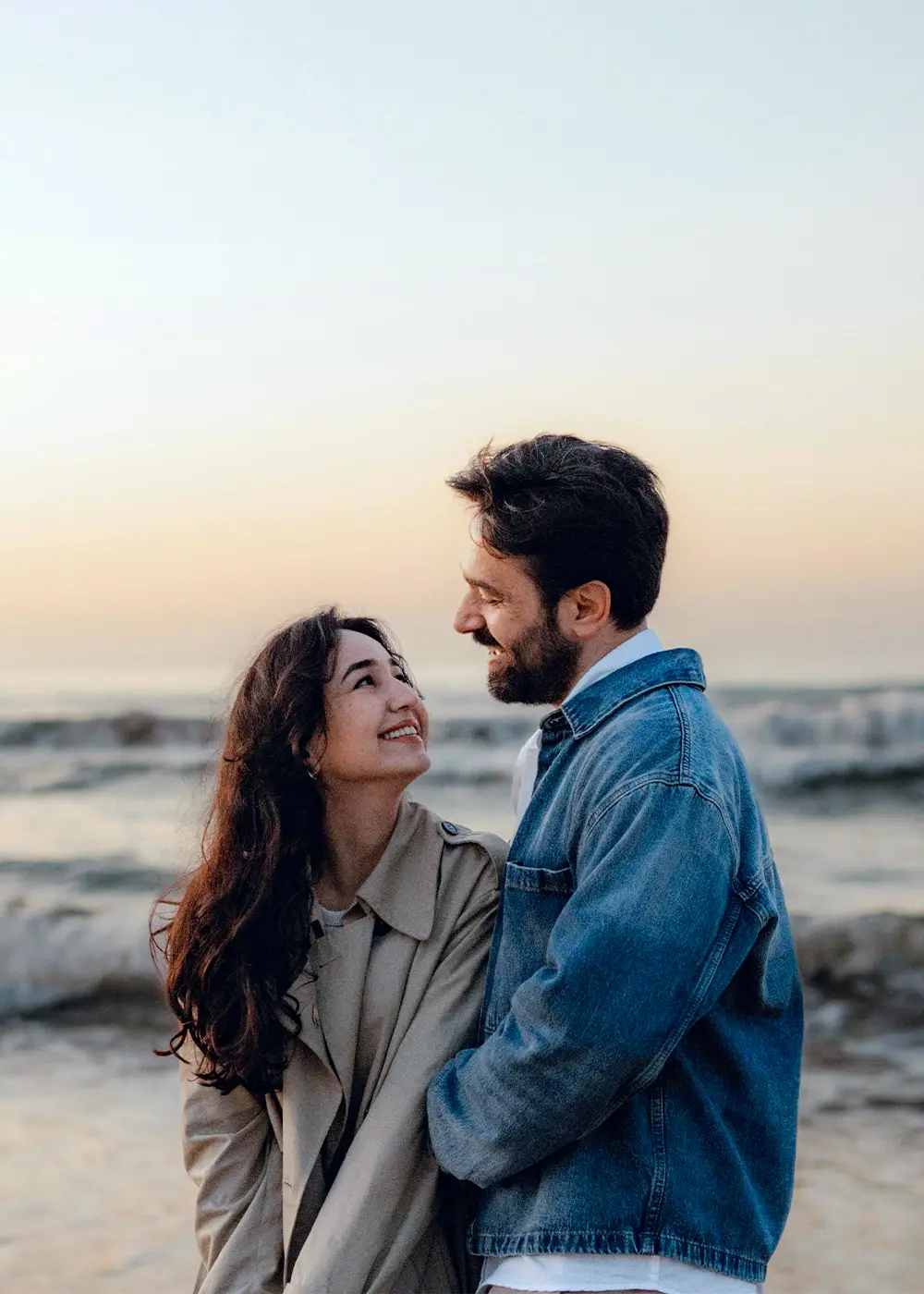Couple smiling and embracing on the beach at sunset with waves in the background.