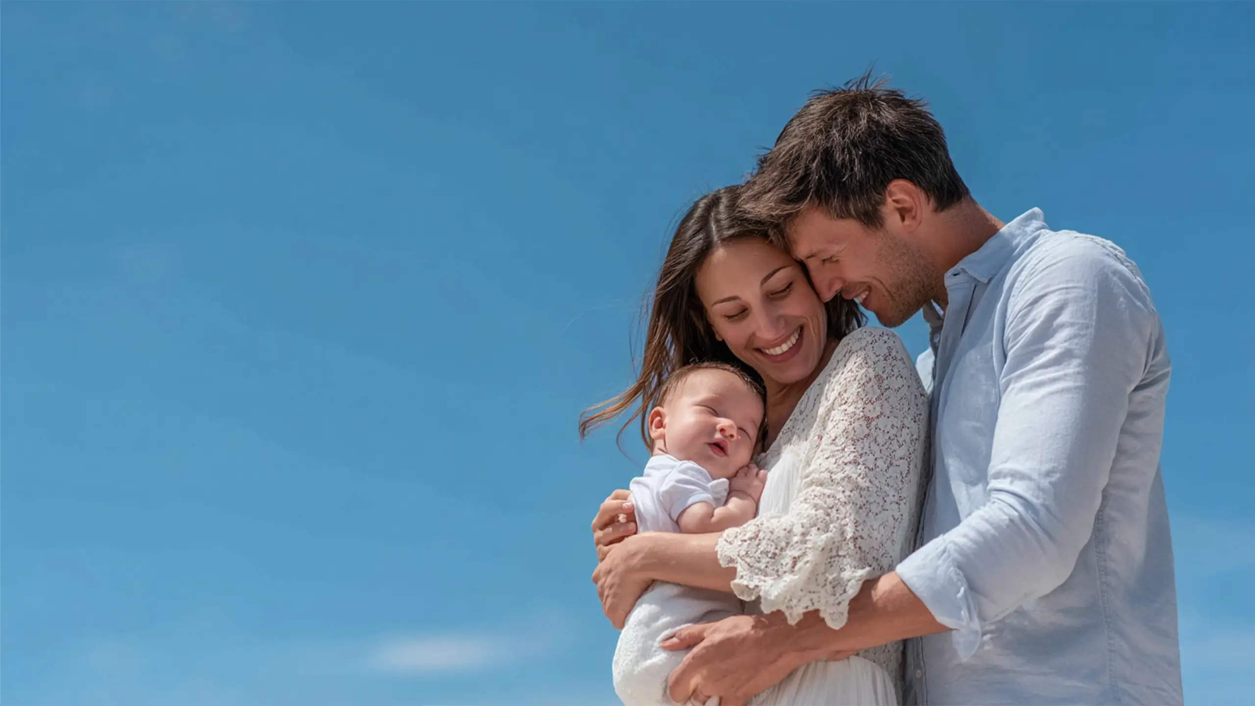 Smiling couple holding a peacefully sleeping baby against a clear blue sky.