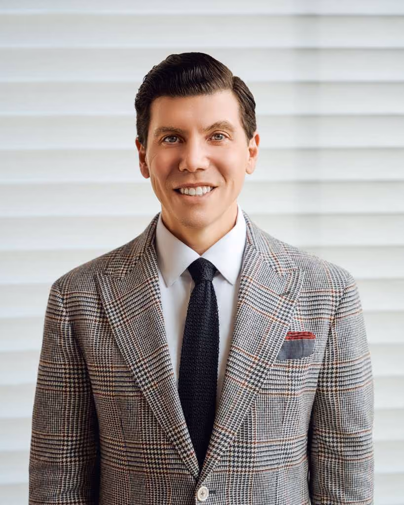 Smiling man with neatly styled dark hair wearing a plaid suit jacket, white dress shirt, black tie, and pocket square in front of white horizontal blinds.