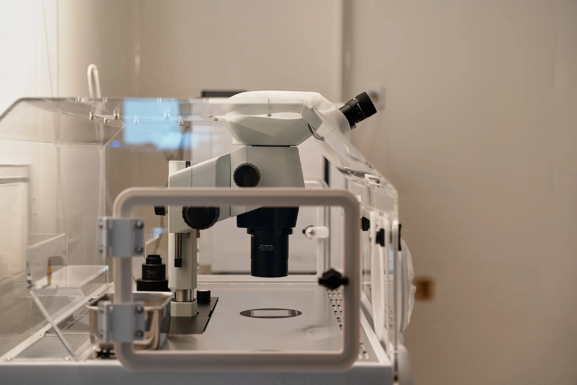 Laboratory microscope enclosed in a clear protective case on a metal table in a clinical setting.