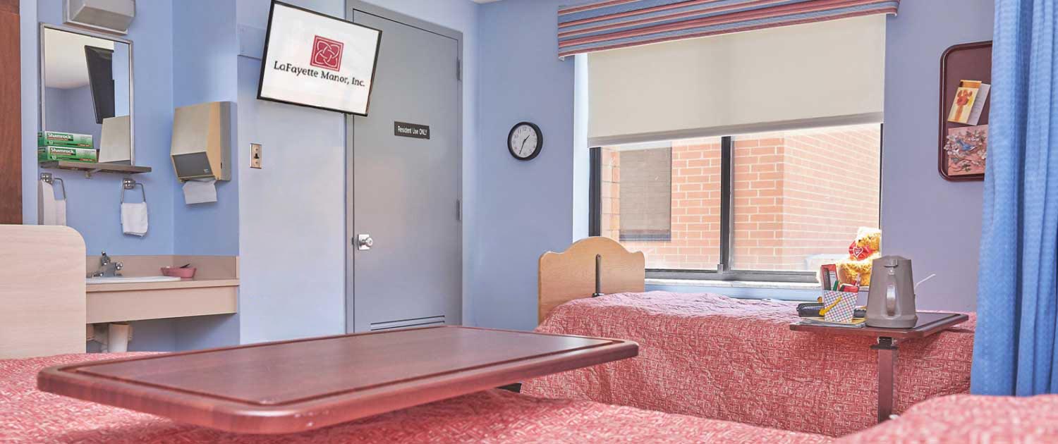 Patient room in medical facility with two beds covered in red blankets, a wall-mounted TV, a sink, and a window with a roller shade.