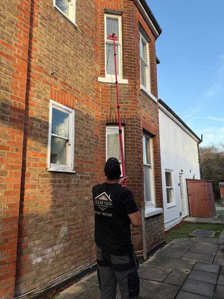 Person wearing a black shirt and cap using an extended pole to clean or reach a second-floor window of a brick house.