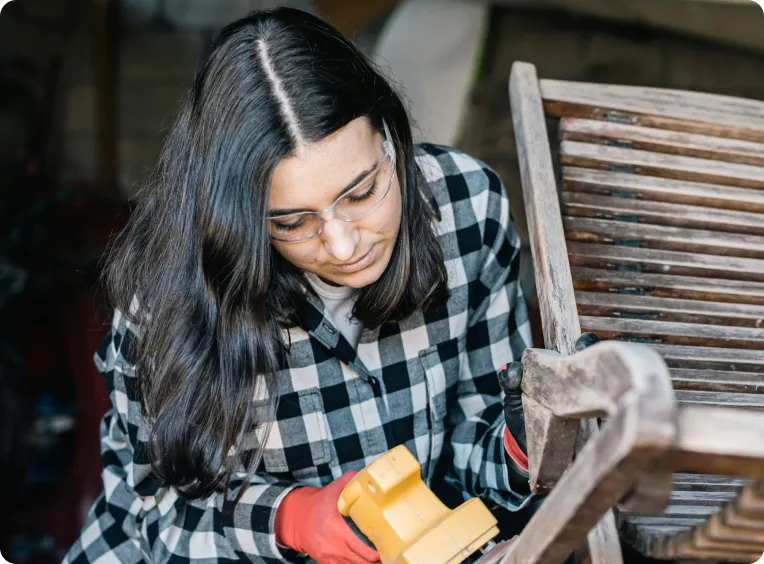 une femme porte des lunettes de protection et ponce sa chaise de jardin en bois