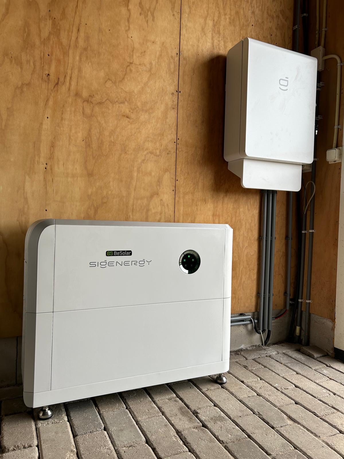 Witte Sigenergy en BeSolar energieopslagapparatuur gemonteerd op een houten muur met kabels en een betegelde vloer.
