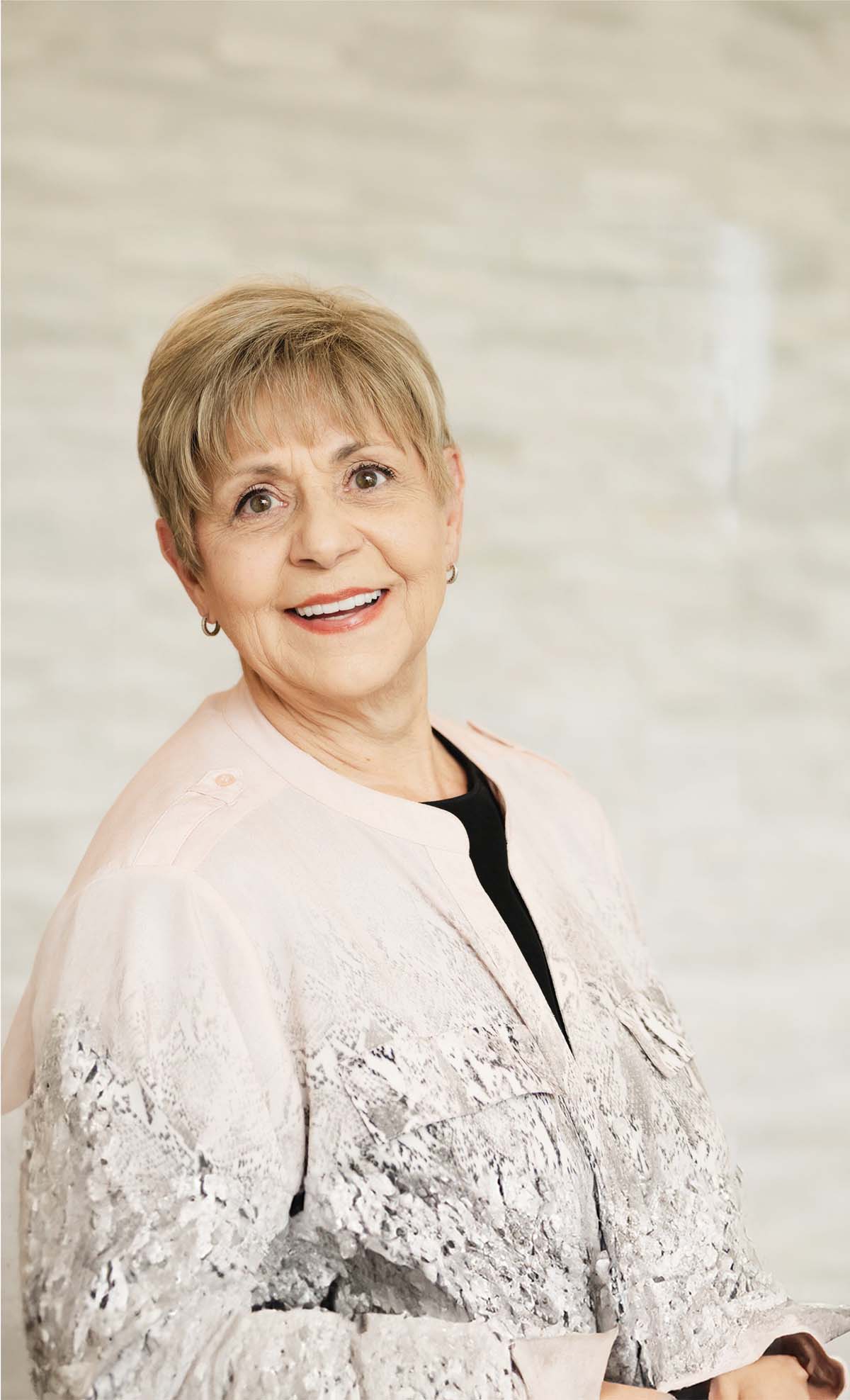 Smiling elderly woman with short blonde hair wearing a light patterned jacket over a black top.