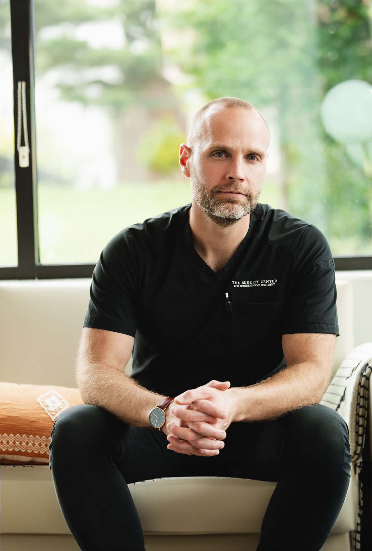 Man with a beard wearing black scrubs sitting on a couch with hands clasped, looking at the camera.