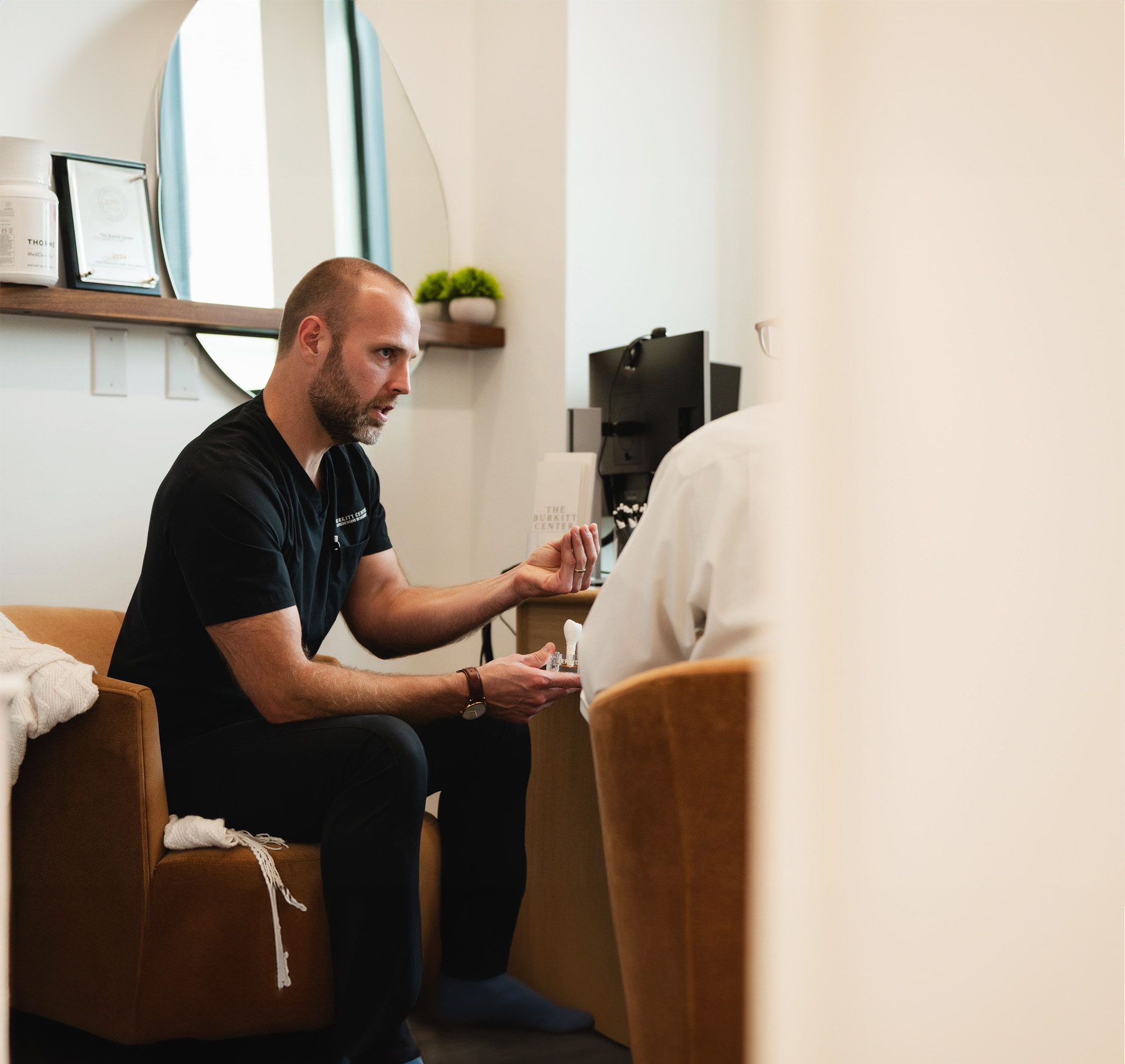 Man wearing black scrubs sitting in a brown chair, explaining or showing a small item to another person in a white shirt in an office setting.