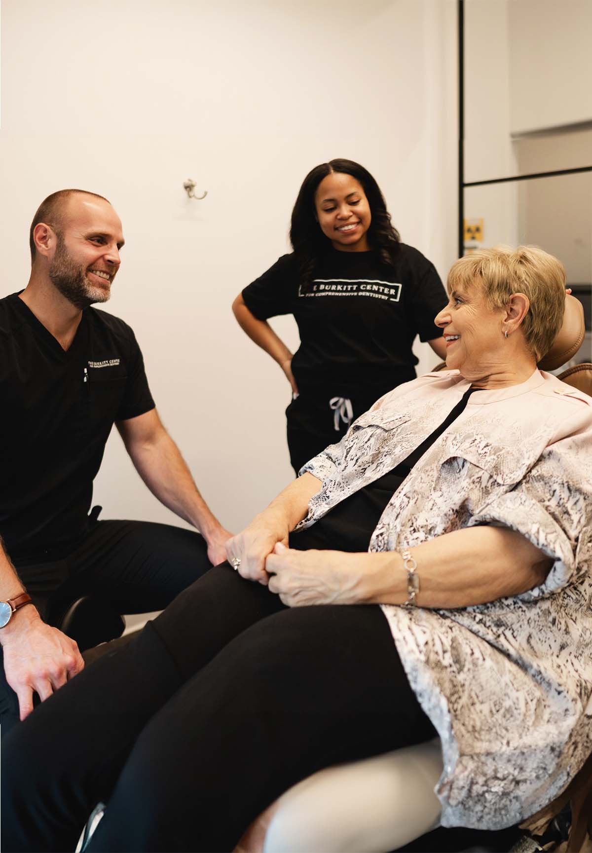 Smiling elderly woman sitting in a dental chair talking with two dental professionals wearing black scrubs.
