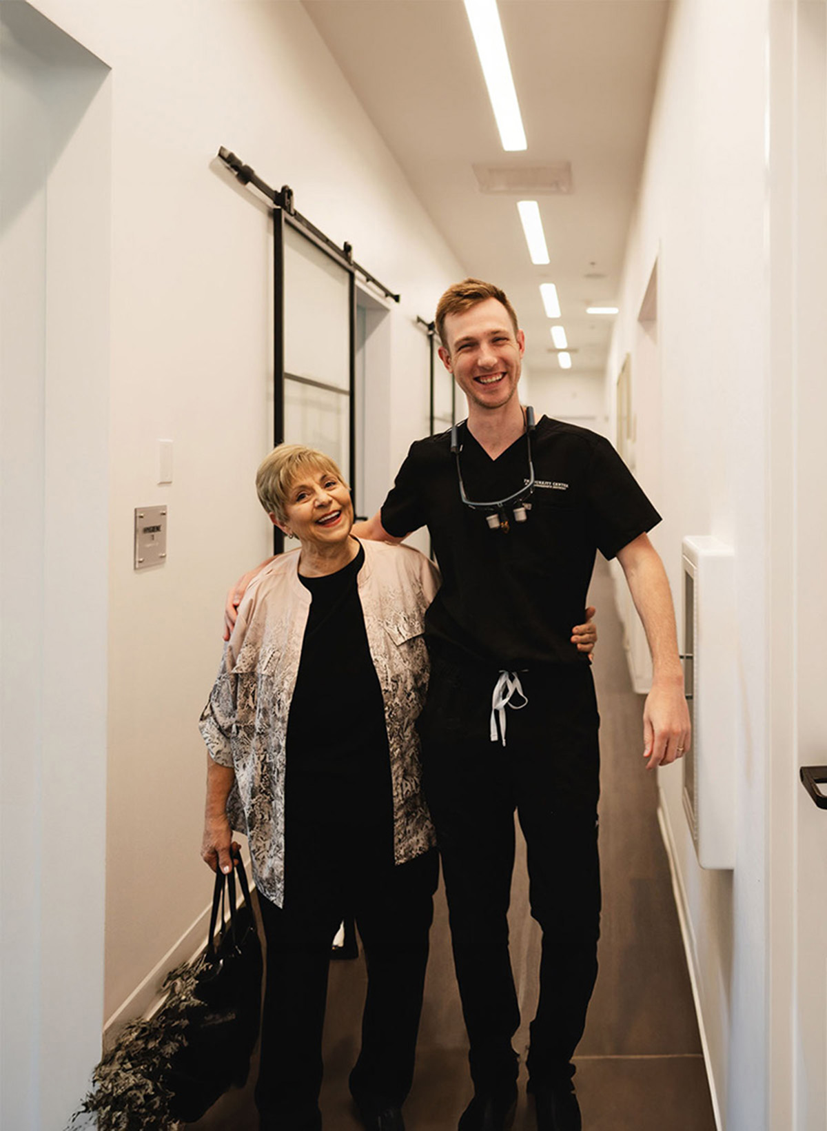 Smiling older woman standing next to a young male healthcare professional in a hallway, both with arms around each other.