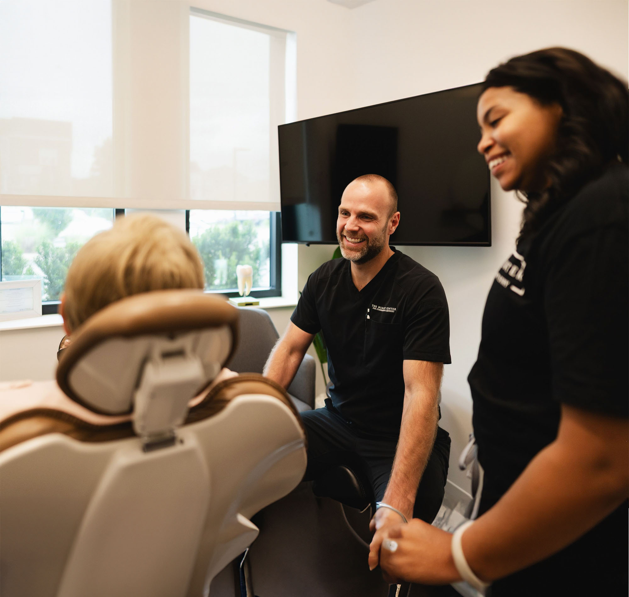 Dentist and dental assistant smiling and talking to a patient seated in a dental chair in a bright office.