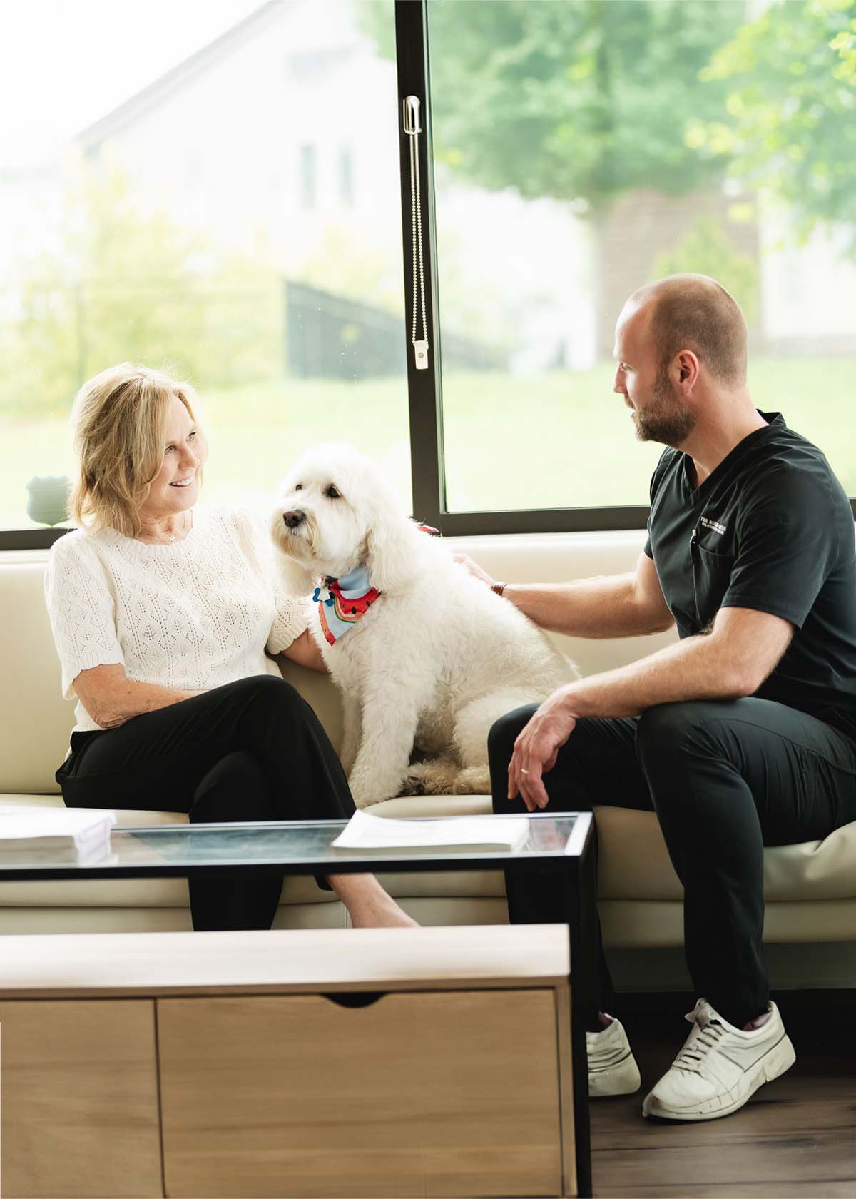 Woman and man sitting on a couch with a white fluffy dog between them, in a bright room with large window.