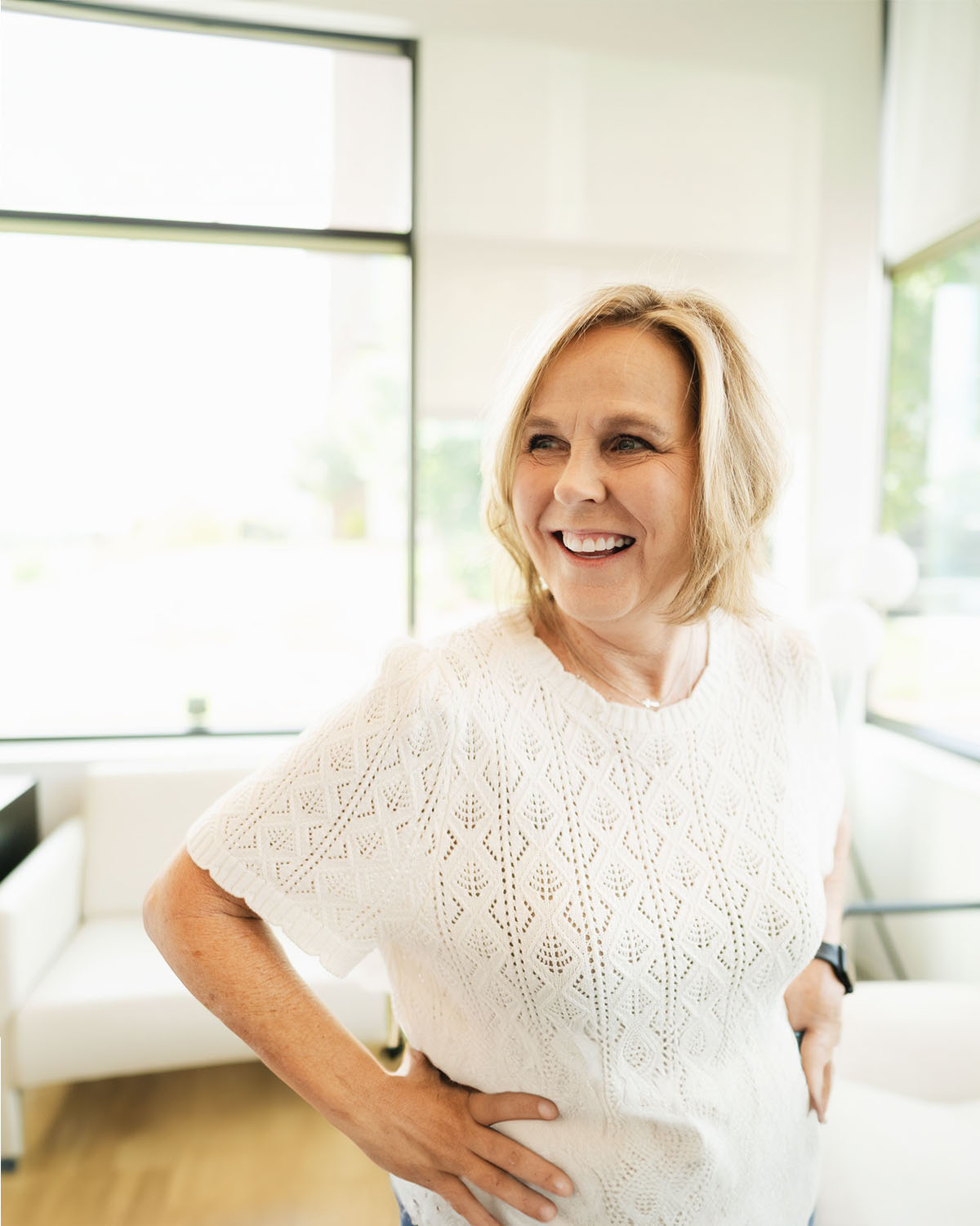 Smiling middle-aged woman with short blonde hair wearing a white knit top standing indoors with hands on hips.