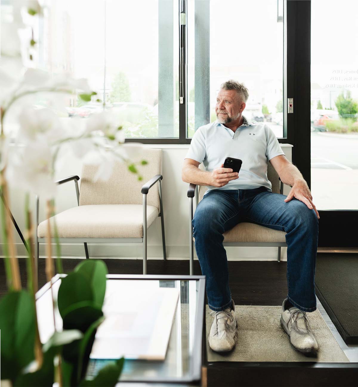 A man in a light blue polo shirt and jeans sitting on a beige chair in a bright waiting area, holding a smartphone and looking out the window.