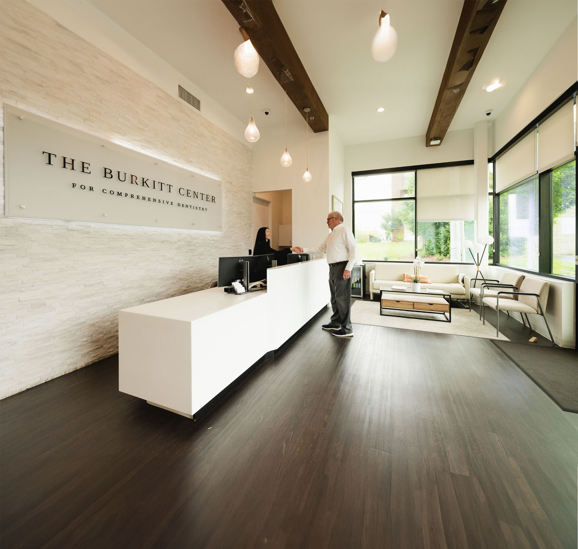Interior of The Burkitt Center for Comprehensive Dentistry reception area with a man and receptionist shaking hands at a white desk.