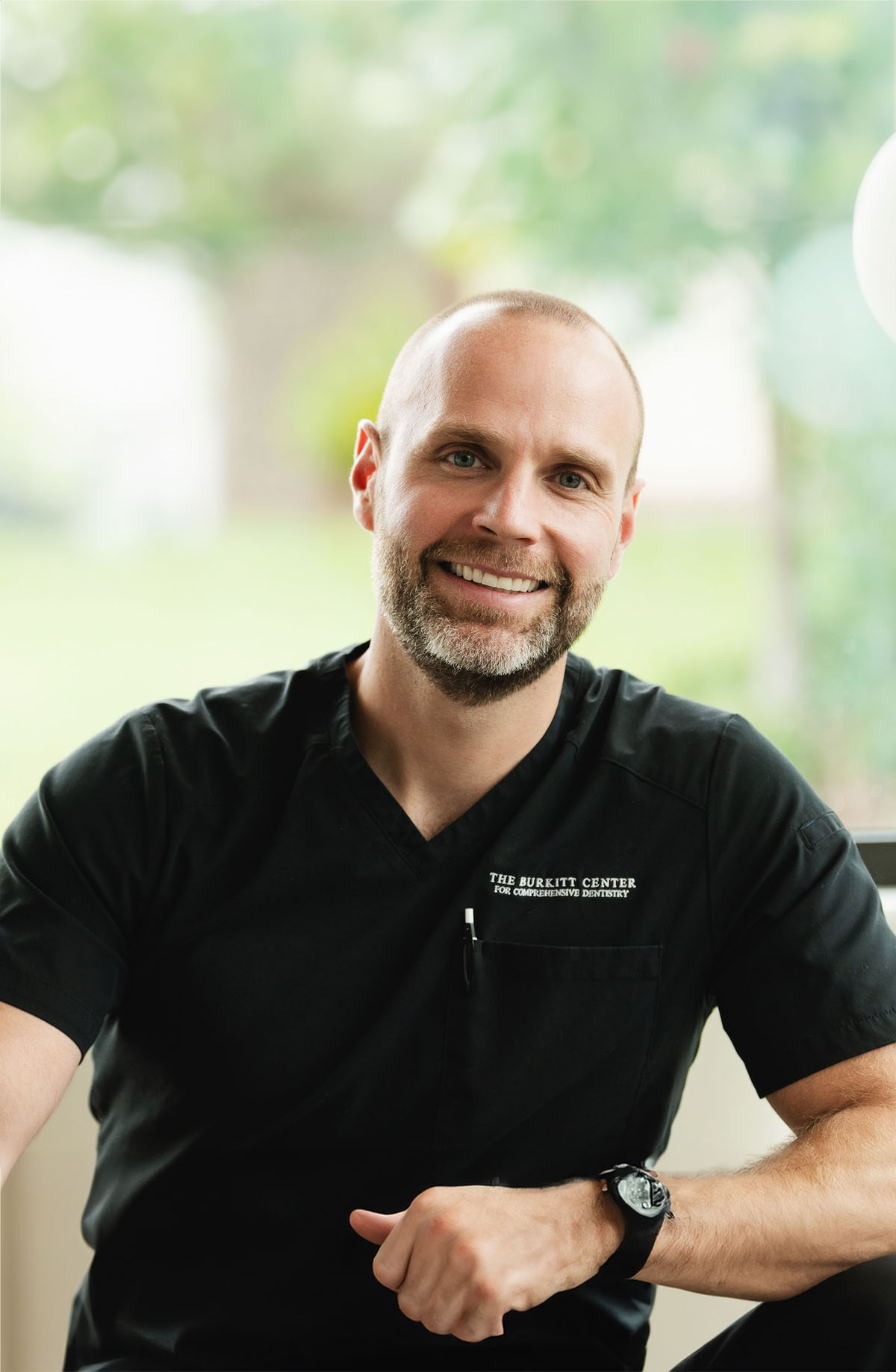 Smiling man in black scrubs with The Burkitt Center for Comprehensive Dentistry logo sitting in front of a window with a blurred green background.