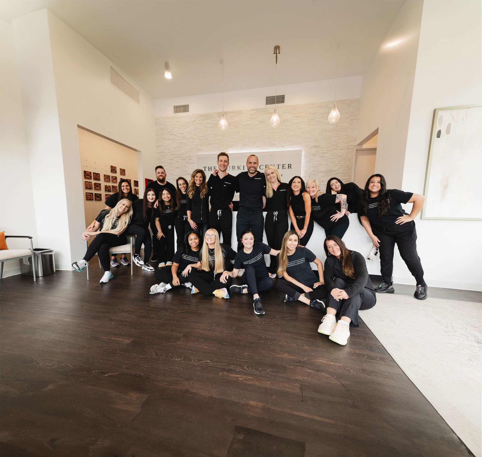 A group of 18 people wearing black uniforms posing together inside a modern office with white walls and wooden floors.