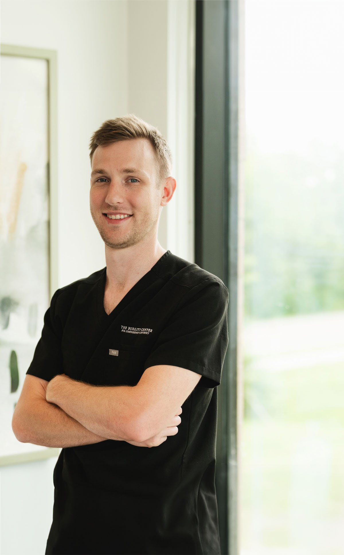 Smiling man in black medical scrubs with arms crossed standing near a window.