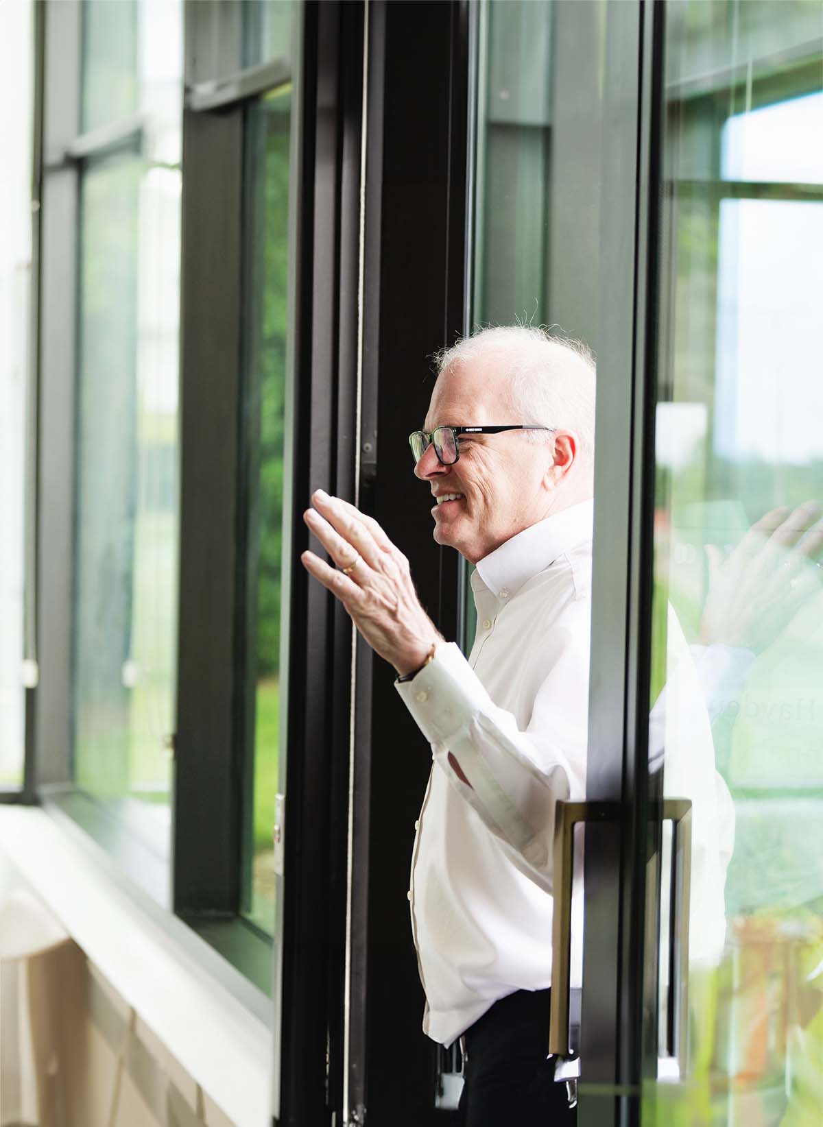 Smiling elderly man in glasses and white shirt waving while standing by a glass door.