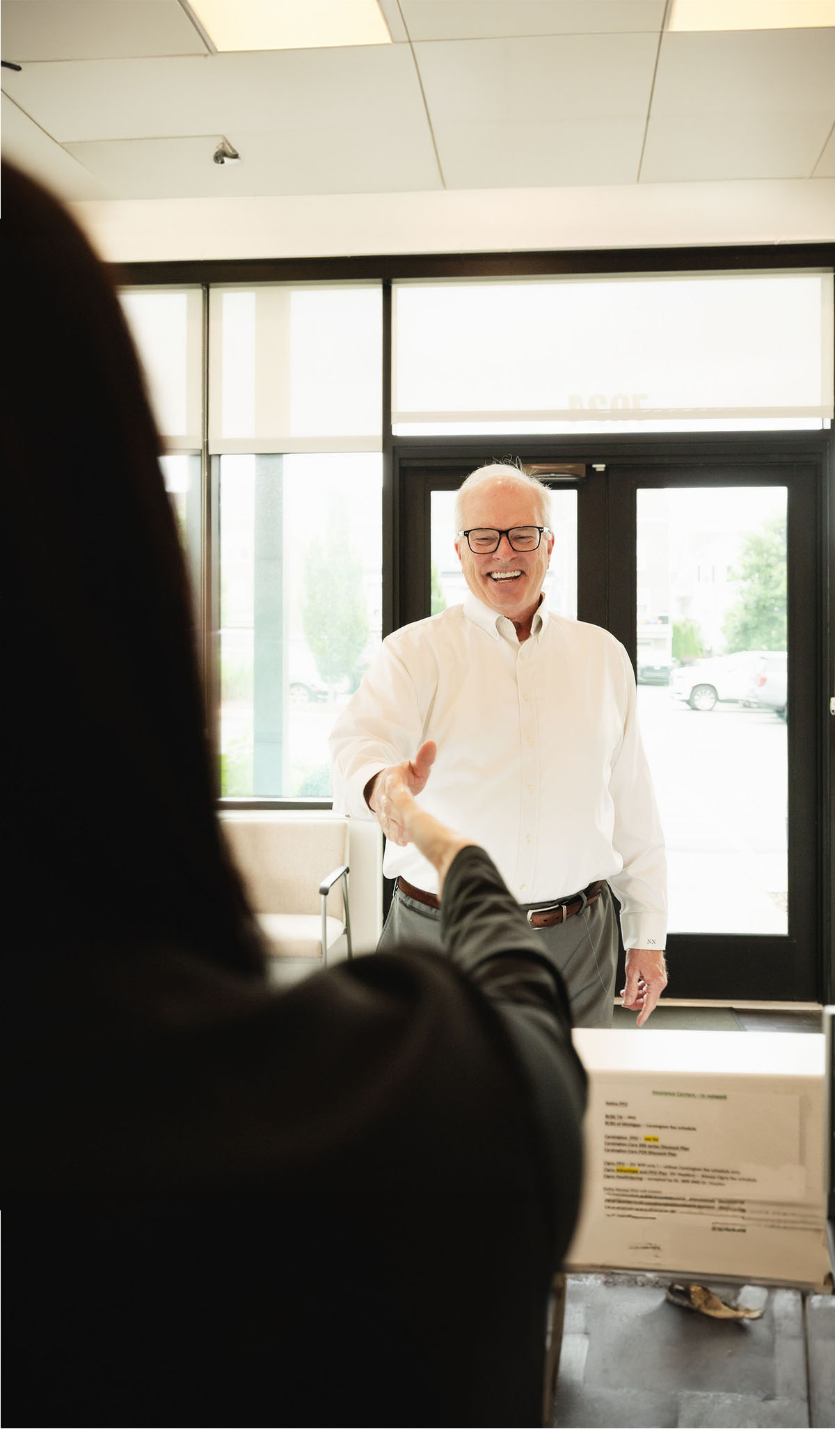 Smiling elderly man wearing glasses and a white shirt reaching out to shake hands inside a bright office lobby.
