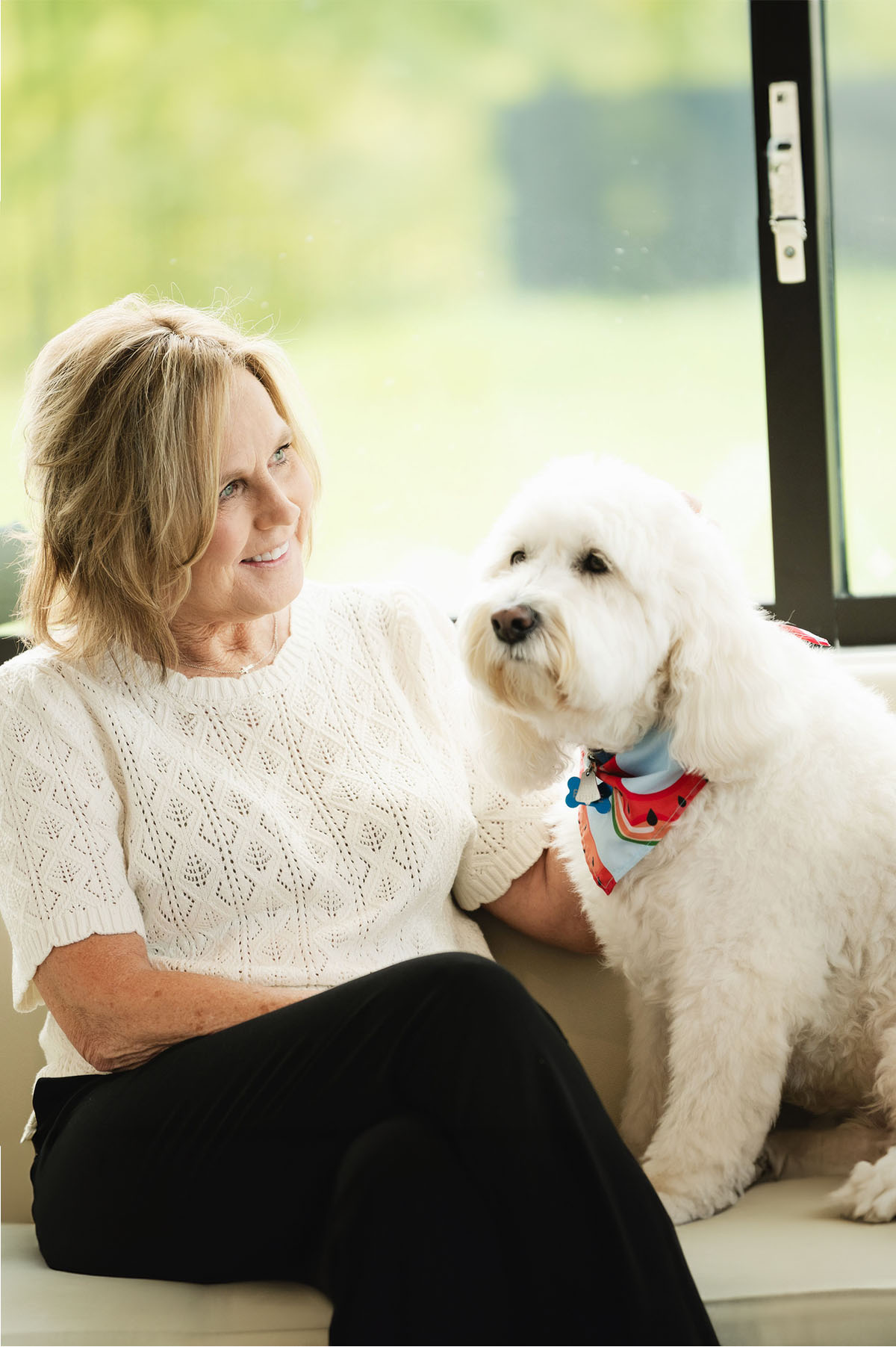 Smiling woman in white sweater sitting on a couch with a fluffy white dog wearing a colorful bandana.