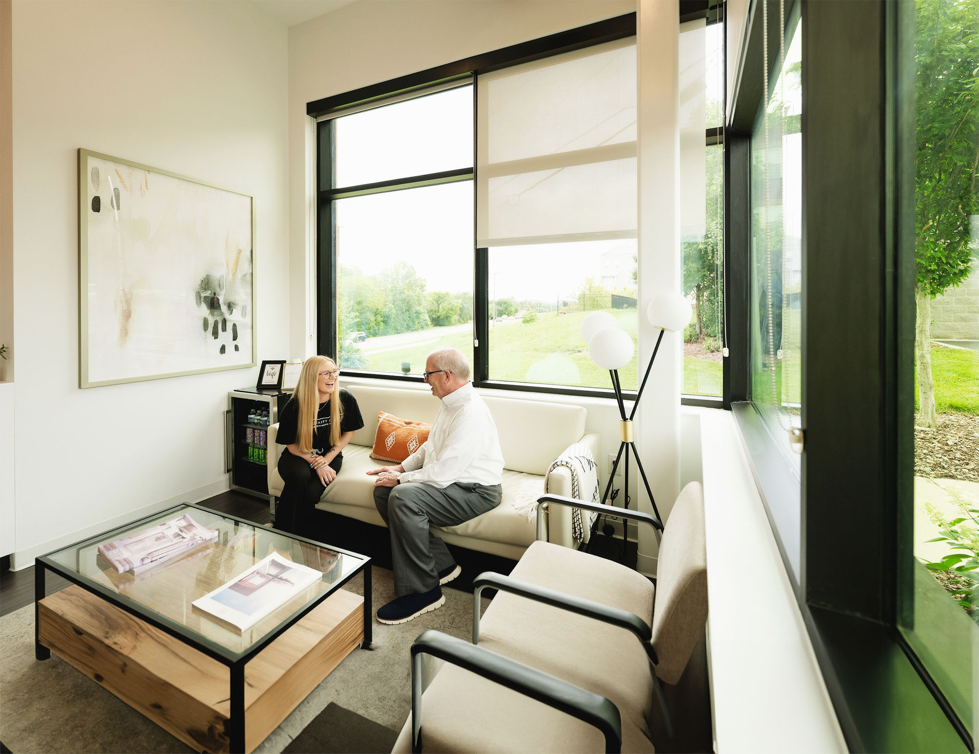 Two people, a woman and an older man, sitting and conversing on a white sofa in a bright room with large windows.