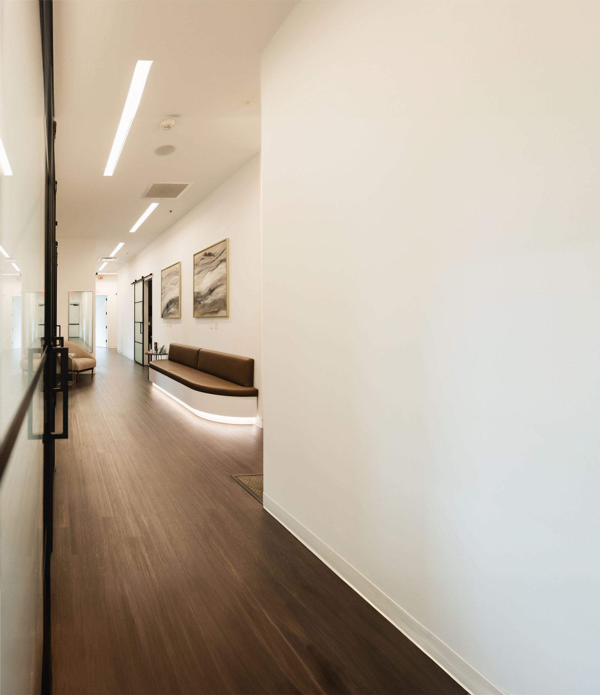 Modern hallway with brown wood flooring, white walls, a brown cushioned bench illuminated underneath, and abstract art on the wall.