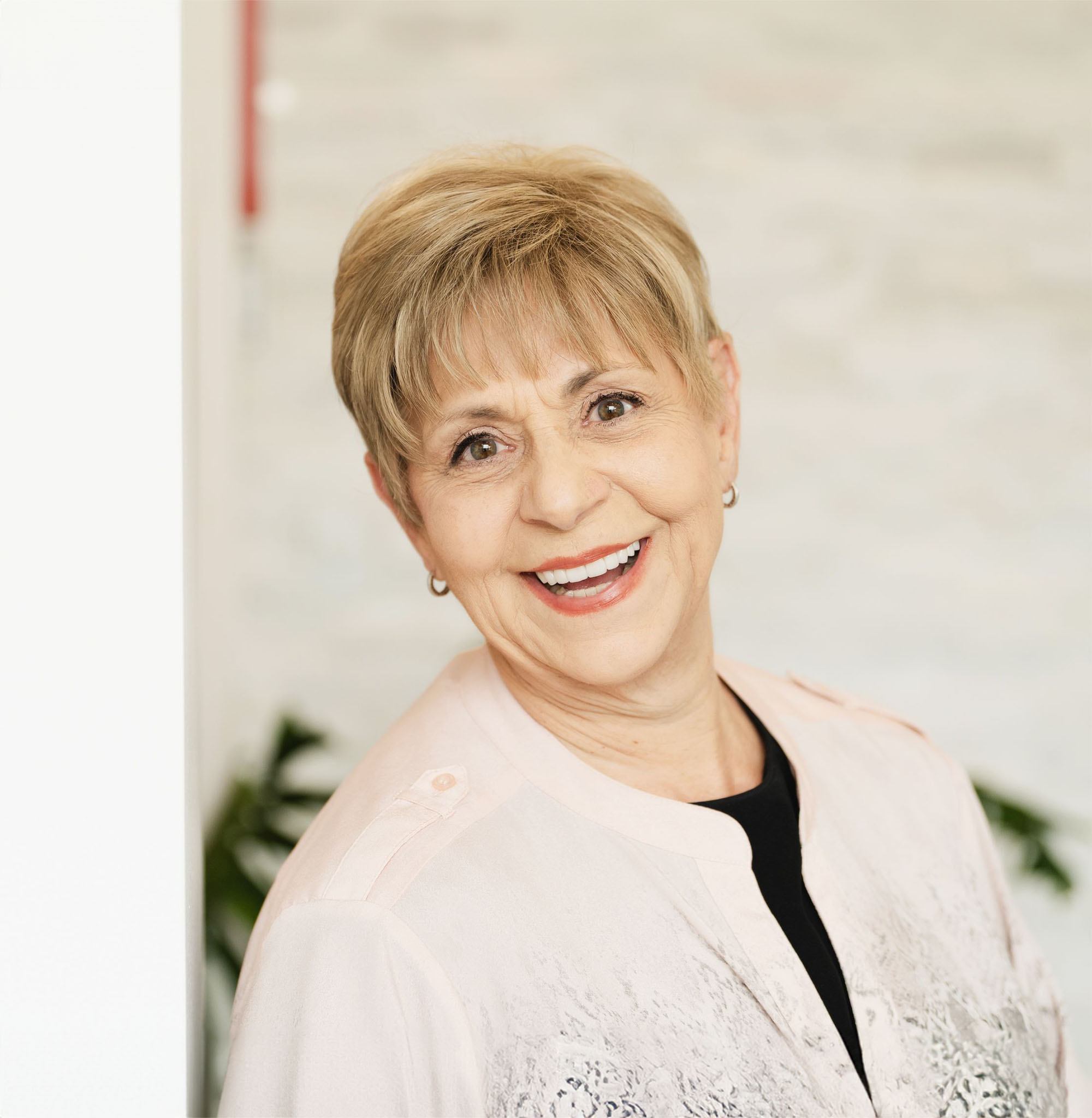Smiling mature woman with short blond hair wearing a light pink blouse and small hoop earrings, standing indoors.