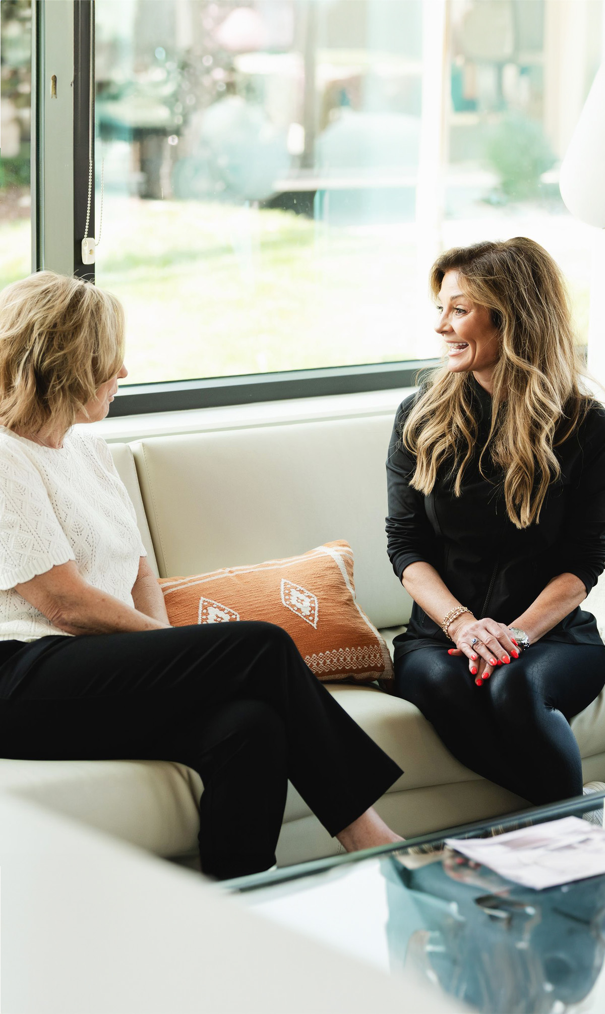 Two women sitting on a beige sofa near a window, engaged in a friendly conversation.