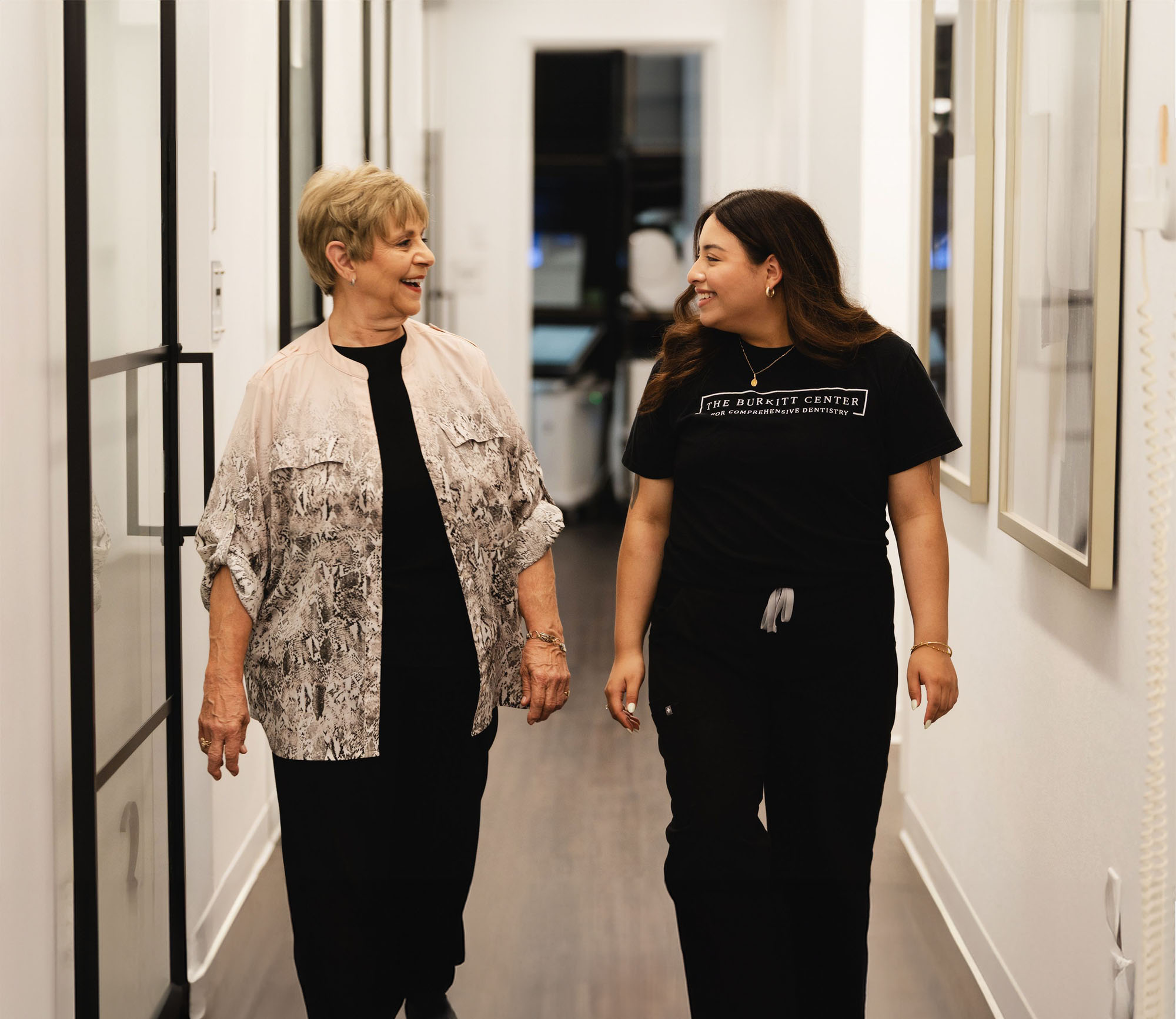 Two women smiling and walking down a hallway, one wearing a black dress with a patterned jacket, the other in black scrubs with The Burkitt Center logo.
