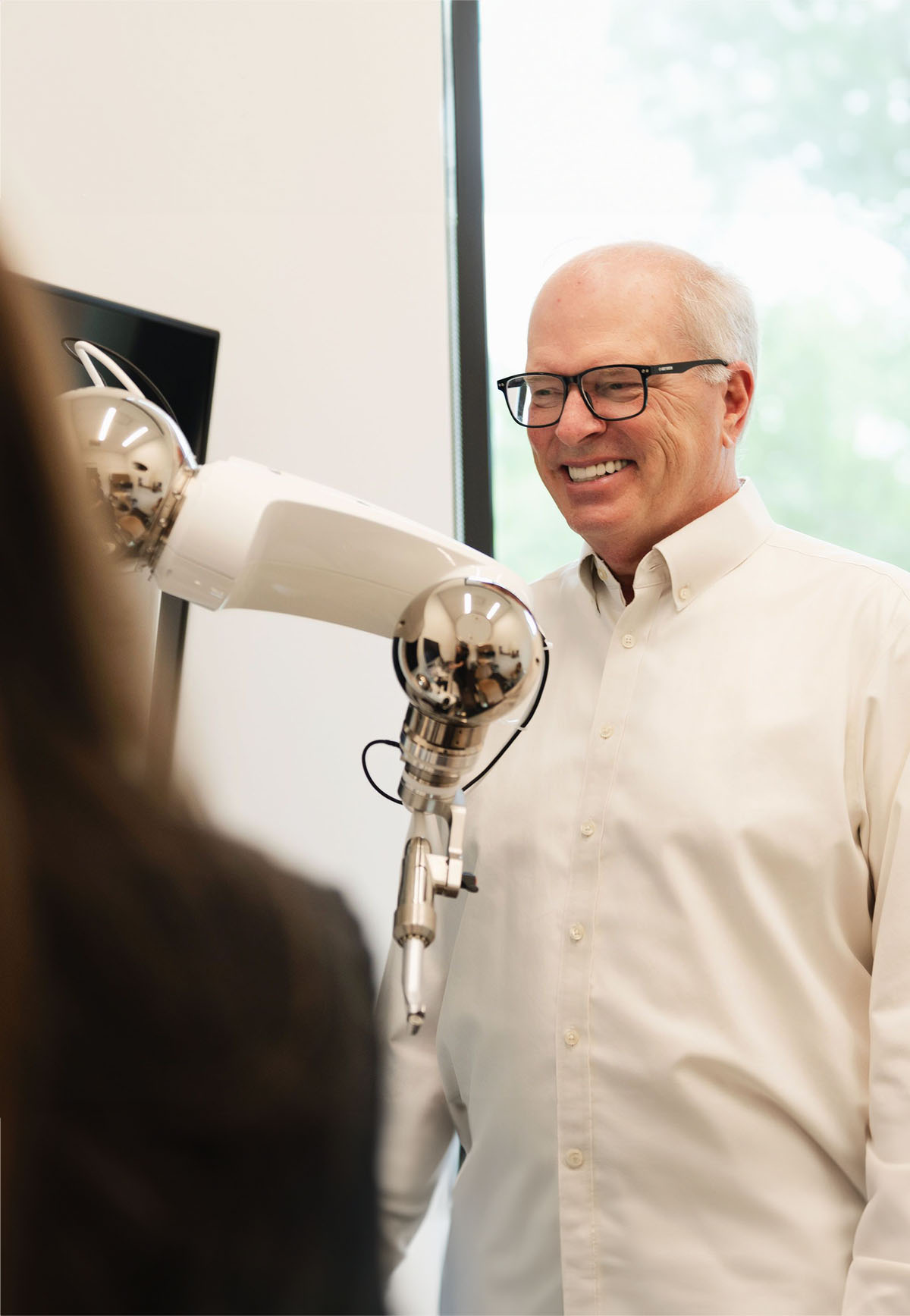 Smiling man in glasses and white shirt interacting with a robotic arm in a modern indoor setting.