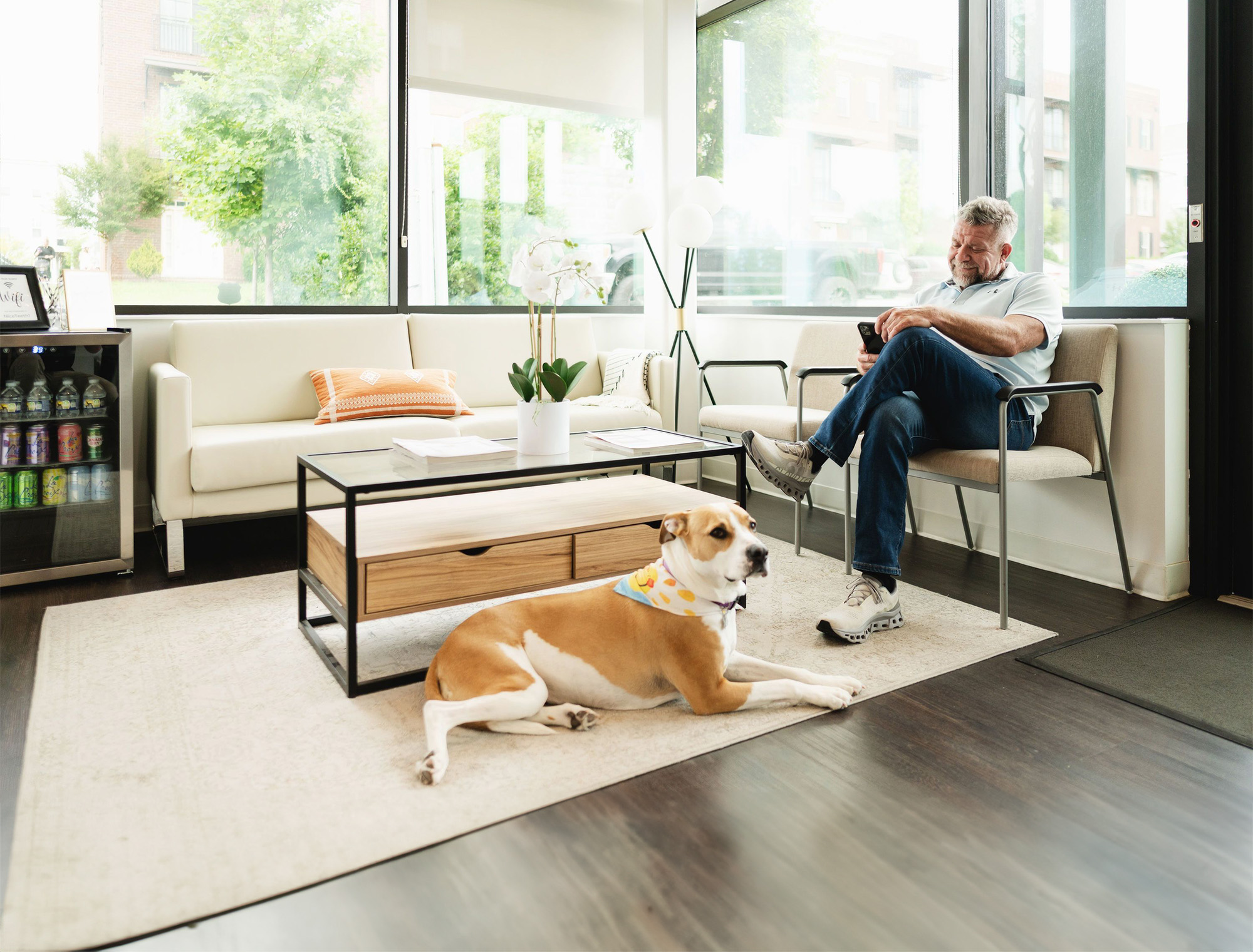 Man sitting on a chair looking at his phone in a bright waiting room with a dog lying on a rug in front of a coffee table.