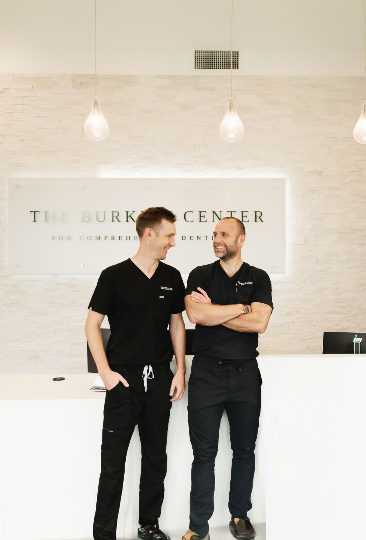 Two male dental professionals in black scrubs standing and smiling in front of a reception desk at The Burkitt Center.