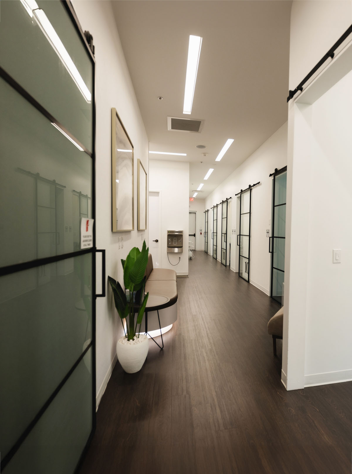 Modern hallway with dark wood flooring, white walls, numbered glass sliding doors, a beige bench, and a potted plant.
