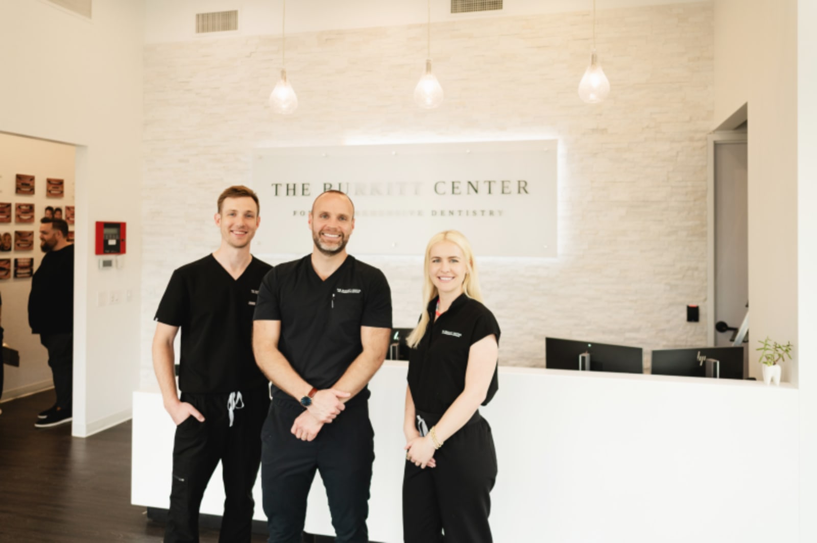 Three dental professionals in black and white scrubs stand together at the front desk of a modern dental office.