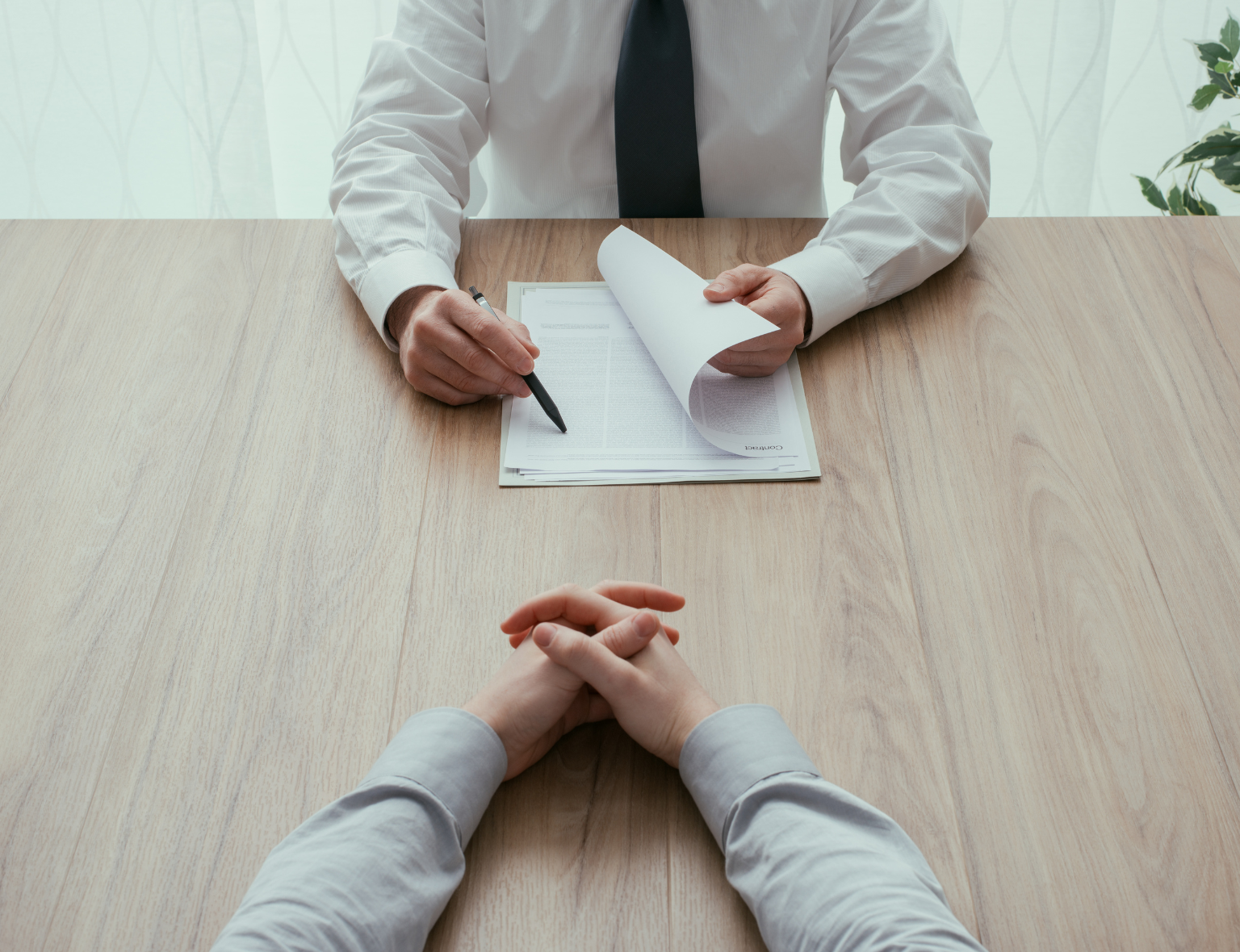 Zwei Personen sitzen sich an einem Holztisch gegenüber, eine davon hält Dokumente und einen Stift, offenbar in einem Gespräch oder Vorstellungsgespräch.