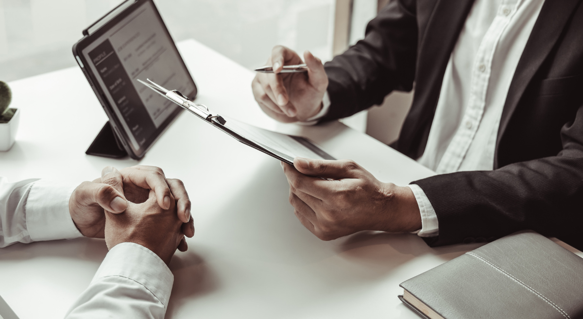 Mehrere Lebensläufe mit einer Brille darauf liegen auf einem Holztisch neben einem Laptop und einer kleinen Uhr.